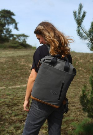 Grauer wasserdichter Rucksack mit braunen Lederelementen, Rolltop-Design, äußere Reißverschlusstasche und verstellbare schwarze Riemen. Außenbereich.