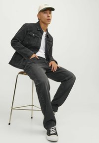 Svart denimjacka och jeans, vit T-shirt, svarta sneakers med vit sula, beige keps. Modell sitter på en kromstol mot en enfärgad bakgrund.