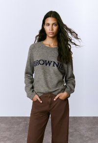 Mujer joven con pelo largo y oscuro, vistiendo un suéter gris con el texto "BROWNIE" y pantalones marrones, con las manos en los bolsillos, de pie contra un fondo sencillo.