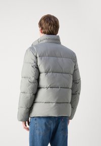 Grå pufferjakke med høj krave, med horisontale syninger og en polstret tekstur. Sammen med blå denim jeans, set fra bageste.