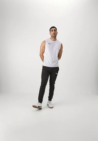 Muž stojící s rukama za zády, oblečený v bílém bezrukávovém tričku Puma, černých joggingových kalhotách Puma a bílých teniskách na jednobarevném bílém pozadí.