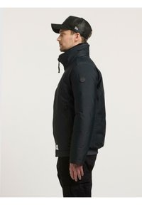 Svart polstret jakke med høy krage, glidelåslommer og en rund logo på ermet; brukt sammen med en svart nettingcaps.