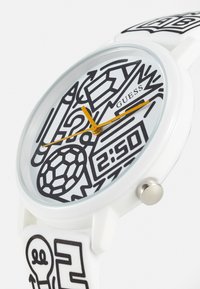 Montre-bracelet Guess blanche avec motifs abstraits noirs, aiguilles jaunes, et un design comprenant une ampoule, un crayon, une carapace de tortue, et une horloge digitale affichant 2:50.