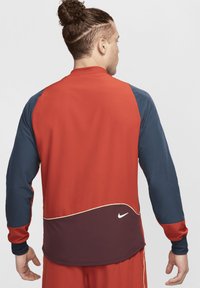 Jachetă sport roșie și bleumarin cu mâneci lungi, textură netedă, tiv curbat și un logo alb proeminent Nike pe spate.