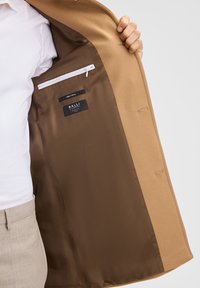 Blazer in lana cammello con foderatura in satin marrone. All'interno è presente una tasca con zip e le etichette del marchio. Presenta cuciture pulite e su misura.