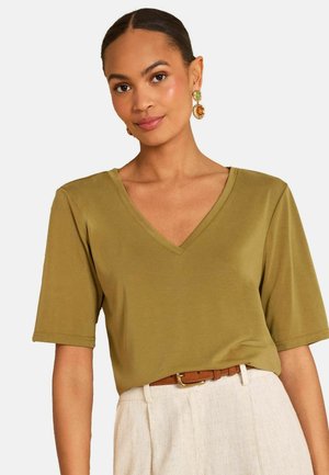 Nainen, jolla on selkään vedetyt hiukset, pukeutuneena oliivinvihreään V-kaula-aukkoinen lyhythihainen paita, beigenväriset housut, ruskea vyö ja vihreä-oranssit roikkuvat korvakorut, katsoo eteenpäin.