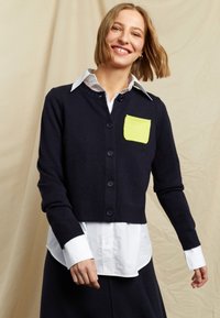 Cardigan a maglia blu navy con un design corto, caratterizzato da una tasca di un vivace verde lime. Ha una chiusura a bottoni e polsini a costine. Sotto indossa una camicia bianca.