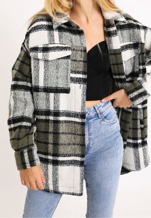 Vrouw draagt een oversized groen, wit en zwart geruite jas over een zwart cropped top en lichtblauwe high-waisted jeans, hand op de heup.