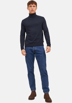 Junger Mann in einem marineblauen Rollkragenpullover, blauen Jeans und braunen Stiefeletten, der vor einem schlichten weißen Hintergrund steht.