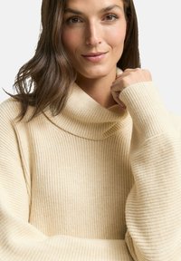 Kobieta z brązowymi włosami, ubrana w sweter z dzianiny w kolorze kremowym z golfem, delikatnie się uśmiecha, trzymając rękę blisko twarzy.