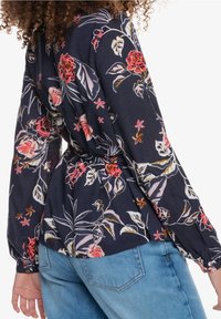Blusa blu navy con vivace motivo floreale in rosso, rosa e verde. Presenta maniche lunghe, vita arricciata e polsini elastici, abbinata a jeans blu.