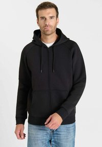 Zwarte zip-up hoodie met een kangoeroezak, verstelbare capuchon met trekkoord en geribbelde manchetten. Gedragen over een wit T-shirt en blauwe spijkerbroek.