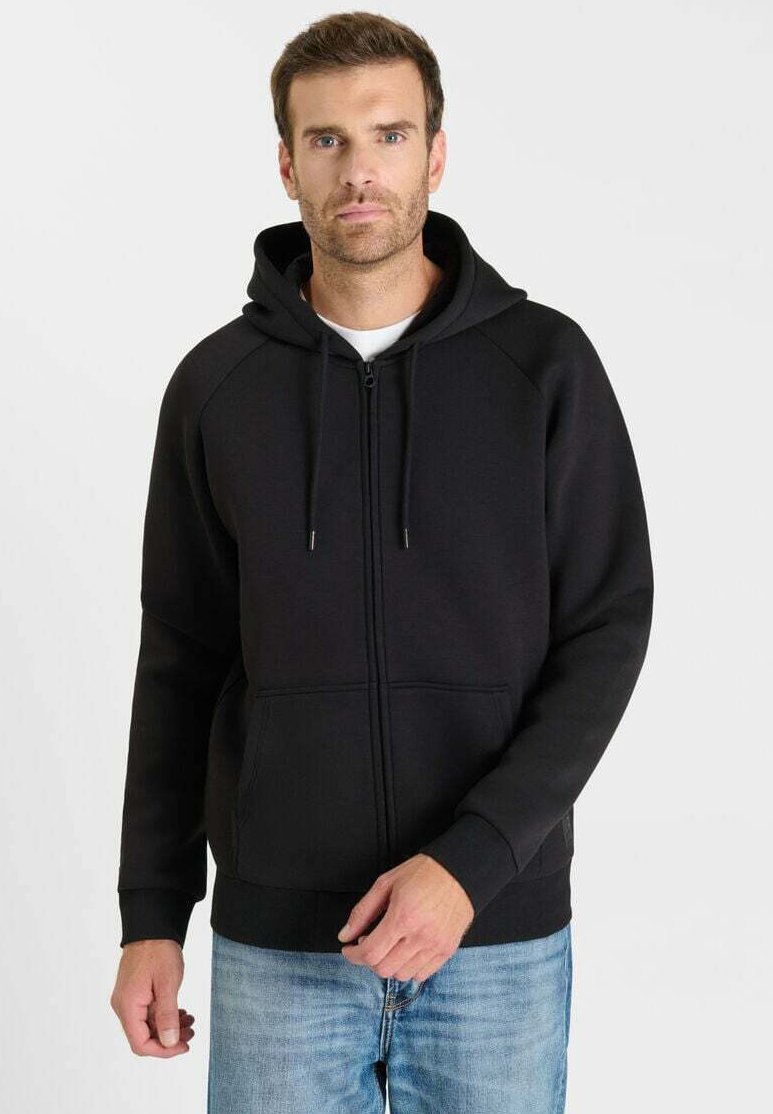 Zwarte zip-up hoodie met een kangoeroezak, verstelbare capuchon met trekkoord en geribbelde manchetten. Gedragen over een wit T-shirt en blauwe spijkerbroek.