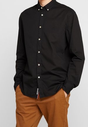 Mand iført en sort skjorte med hvide knapper og brune bukser, hænderne i lommerne, på en ensfarvet grå baggrund.