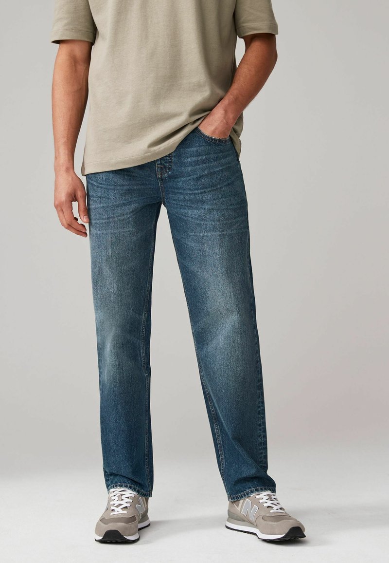 Vaqueros de mezclilla azul con corte recto, lavado medio, desvanecido sutil y diseño de cinco bolsillos, combinados con zapatillas grises y blancas.