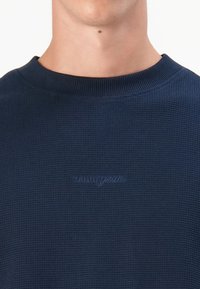Marineblauwe wafelgebreide trui met een ronde halslijn en geborduurd logo op de borst. Het materiaal lijkt textuur te hebben en zacht te zijn.