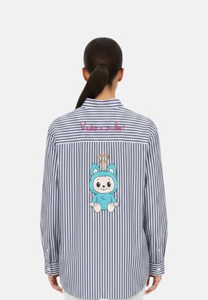 Donna con capelli scuri legati in una coda di cavallo indossa una camicia a righe blu e bianche con un animale blu in stile cartone animato e testo rosso sulla schiena.