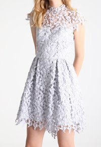 Harmaa mekko korkealla kaula-aukolla, jossa on lehtimäisten kaiverrusten teksturoitu päällinen ja liehuva helma, joka ulottuu polvien ylle.