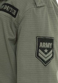 Veste militaire olive avec un tissu texturé. Présente des patchs brodés noirs : "DUTCH" sur une épaule et "ARMY" avec des chevrons sur l'autre.