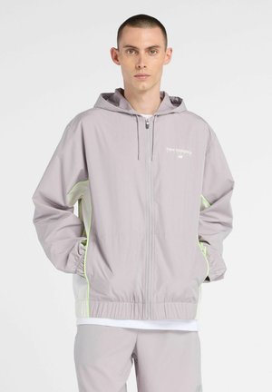 Jonge man draagt een lichtgrijze zip-up capuchonjas met neongele rand en bijpassende broek, staand tegen een effen achtergrond.