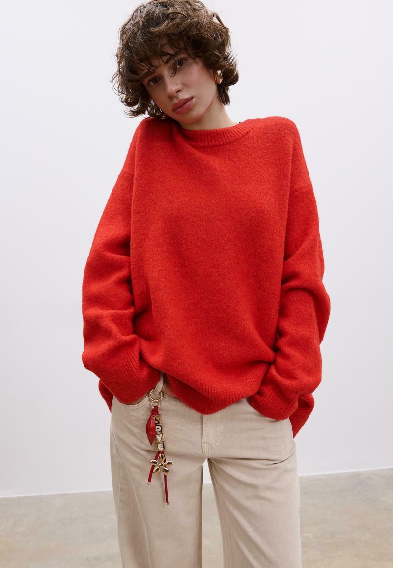 Ein locker sitzender, leuchtend roter Pullover mit einem gerippten Halsausschnitt, kombiniert mit cremefarbenen Hosen, die mit einem dekorativen Schlüsselanhänger mit goldenen Akzenten versehen sind.