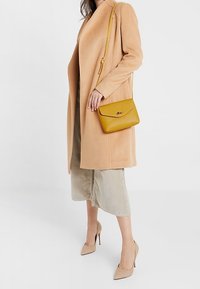 Cappotto di lana beige, lunghezza ginocchio. Gonna grigia chiara, al polpaccio. Borsa a tracolla gialla in pelle con chiusura, e tacchi alti color nude.
