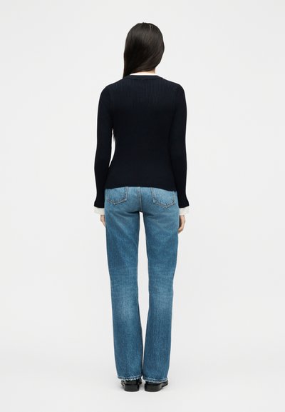 Γυναίκα που στέκεται με την πλάτη προς τα εμπρός, φοράει μαύρο ριμπ μακρυμάνικο μπλουζάκι, μπλε τζιν με ίσια γραμμή και μαύρα παπούτσια σε λευκό φόντο.