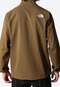 Kastanjebruine softshell jas met een hoge kraag, elastische manchetten en een wit logo op de achterkant. Heeft een gladde textuur en een getailleerde pasvorm.