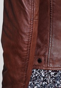Ärmel und unterer Rückennäht einer braunen Lederjacke mit einem schwarzen Druckknopf über einem Rock oder Kleid mit Blumenmuster.