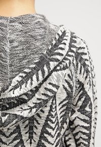 Nærbillede af sort-hvidt strikket stof med bladmotiv på en hættetrøje båret af en person.