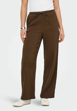Femme portant un pantalon large marron texturé avec taille à cordon, baskets blanches, haut blanc et bracelet manchette argenté.