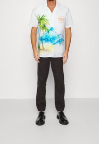 Camisa blanca de manga corta con vibrantes palmeras y montañas en acuarela, diseño de botones, combinada con pantalones negros a rayas y zapatos negros.