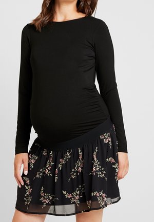 Kobieta w ciąży ubrana w dopasowaną czarną bluzkę z długim rękawem i czarną spódnicę w kwiaty w różowe i zielone wzory.