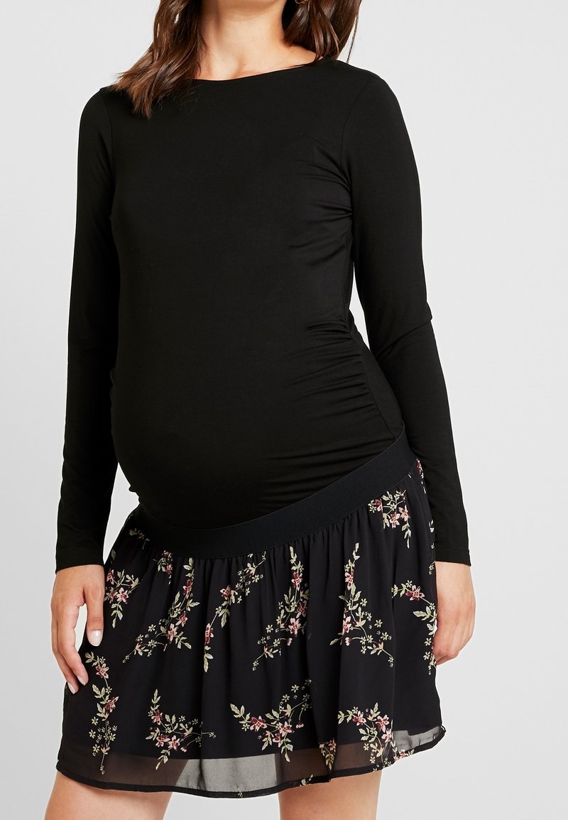 Femme enceinte portant un haut noir ajusté à manches longues et une jupe noire à motifs floraux roses et verts.