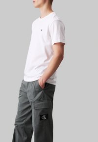 Witte katoenen T-shirt met een klein zwart logo op de linkerkant van de borst, gecombineerd met grijze cargobroek met een patchzak en logo-detail.