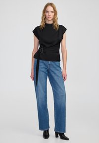 Donna che indossa un top nero a maniche corte con nodo laterale, jeans blu a gamba dritta e stivali neri con tacco alla caviglia su uno sfondo grigio uniforme.