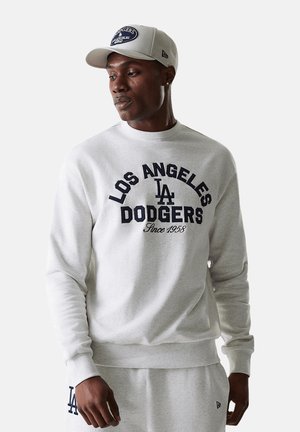 Ein Mann trägt einen hellgrauen Los Angeles Dodgers Sweatshirt, passende Jogginghose und eine Baseballkappe mit Teamlogos.