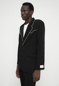 Blazer preto com textura de bolinhas, lapelas de recorte e costura branca contrastante; apresenta dois bolsos frontais e uma etiqueta na manga.