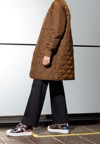 Brun kviltad lång kappa med avslappnad passform, tillsammans med svarta vida byxor och flerfärgade mönstrade sneakers med vit sula.