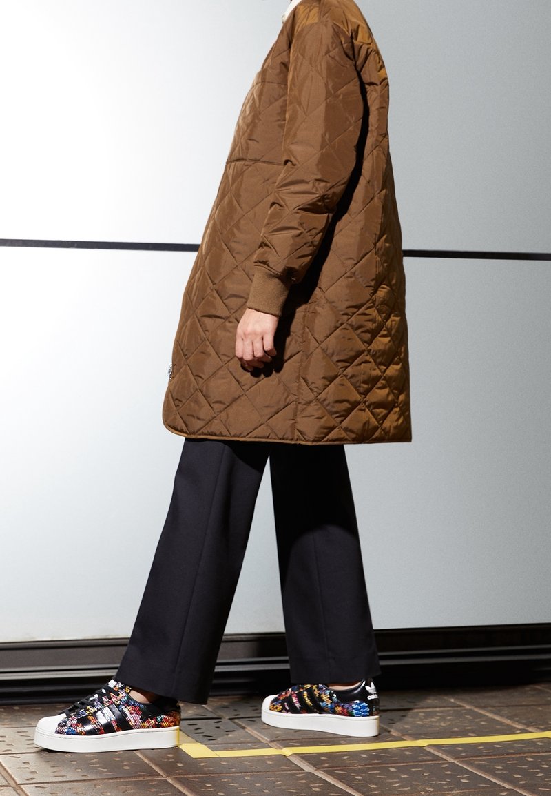 Brun kviltad lång kappa med avslappnad passform, tillsammans med svarta vida byxor och flerfärgade mönstrade sneakers med vit sula.