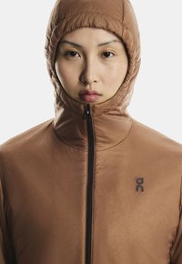 O persoană îmbrăcată într-o jachetă cu glugă de culoare maro, cu fermoarul închis complet, arătând un prim-plan al feței și toracelui superior cu un logo negru pe piept.