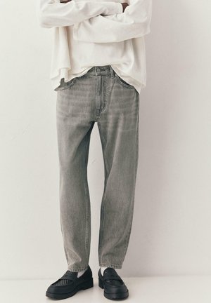 Persoon draagt lichtgrijze spijkerbroek, zwarte instapschoenen, witte sokken en een wit shirt met lange mouwen en gekruiste armen.