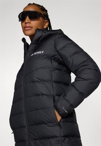 Black puffer jacket with a zippered front, quilted texture, and logo detail on the chest. Features a stand collar and side pockets.