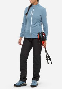 Veste en polaire bleu clair avec fermeture éclair blanche, pantalon de randonnée noir et chaussures de randonnée bleues. Tient des bâtons de trekking avec des accents rouges.