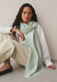 Mujer con pelo largo y oscuro sentada en el suelo, lleva pantalones beige, camisa blanca, zapatos planos negros y una bufanda verde claro con motivos de hojas doradas.
