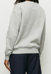 Sudadera gris con un ajuste relajado, puños y dobladillo acanalados, y un cuello redondo. Tela suave con una textura lisa. Combinada con pantalones de color oscuro.