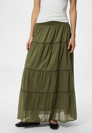 Kvinne iført olivengrønn lagdelt maximalang skjørt med blonder, hvit ermeløs topp og svarte ballettsko, stående på hvit bakgrunn.