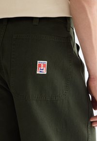 Pantalon vert avec une poche arrière carrée avec un petit patch logo Wrangler, porté par une personne avec une chemise beige et un bras visible.
