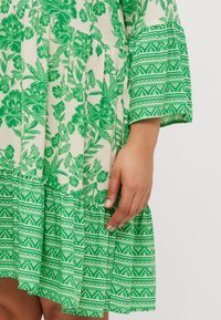 Πράσινο φλοράλ φόρεμα με υφασμάτινη υφή, διαθέτοντας στρώσεις ραμμένες στις άκρες, μακριές καμπάνες μανίκια και αντίθετα μοτίβα σε ελαφρύτερη απόχρωση.