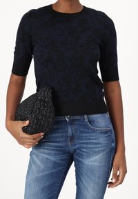 Marinblå kortärmad tröja med ett svart blommönster, kombinerad med blå denimjeans och en texturerad svart vävd väska.